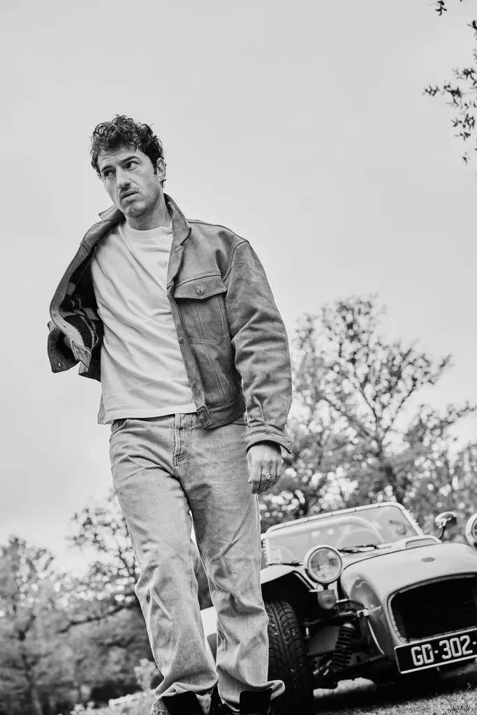 A man in a jacket and jeans walking toward the camera in front of a vintage sports car, in black and white.
