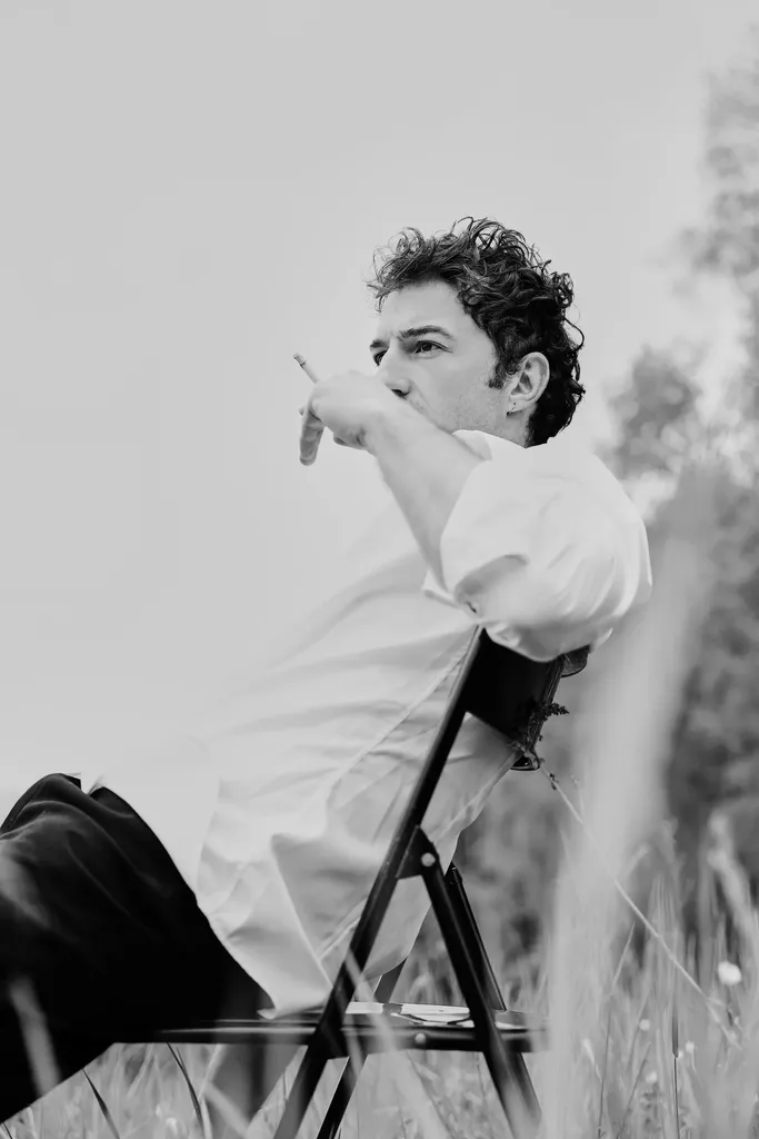 A man in a white shirt sitting on a folding chair in the grass, one arm outstretched, in black and white.