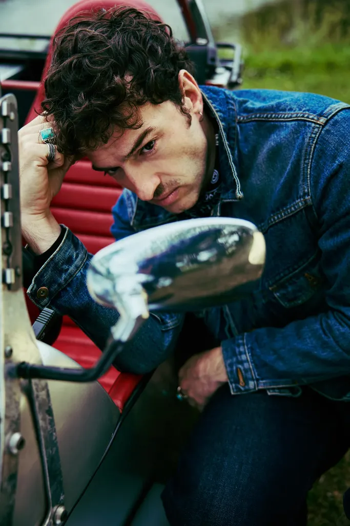 A man in a denim jacket leaning on the steering wheel of a vintage car with red seats, colour.