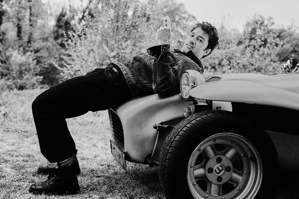 A man lying back on the bonnet of a vintage car in a garden, horizontal frame, in black and white.