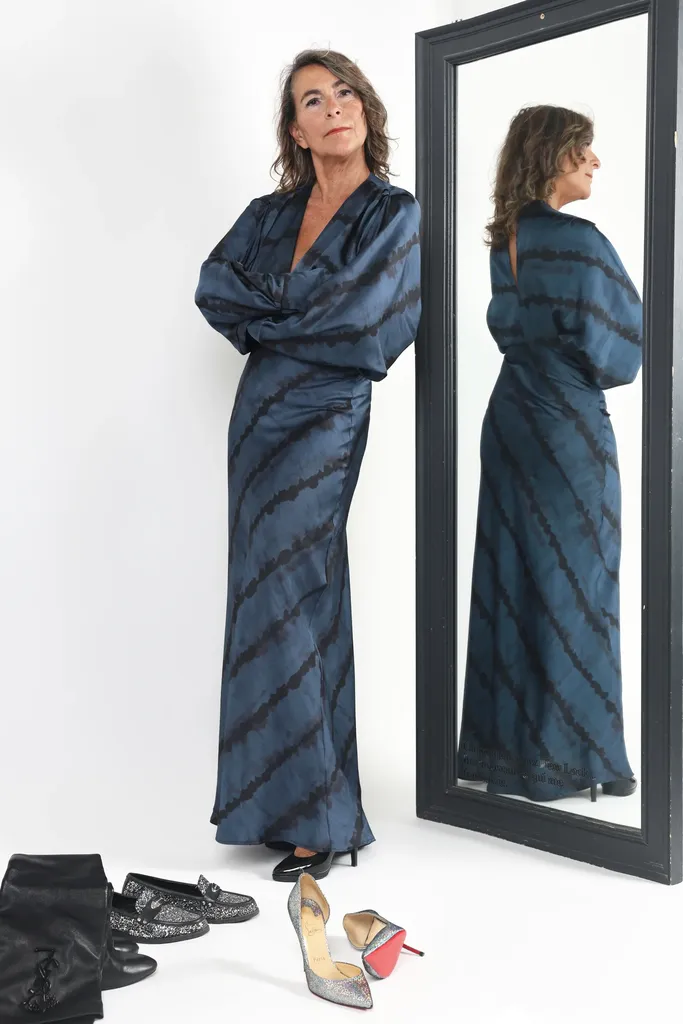 A woman in a long striped dress standing by a tall mirror that reflects her, shoes on the floor, white studio.
