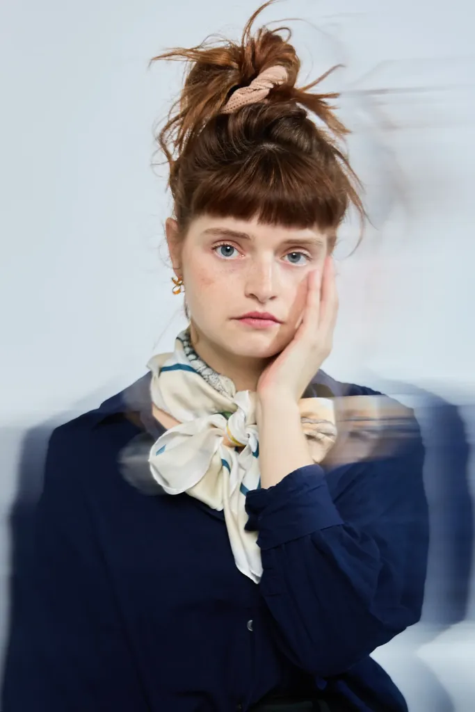 Portrait of a red-haired woman in a navy jumper and pale scarf, hand on her cheek, grey-blue ground.