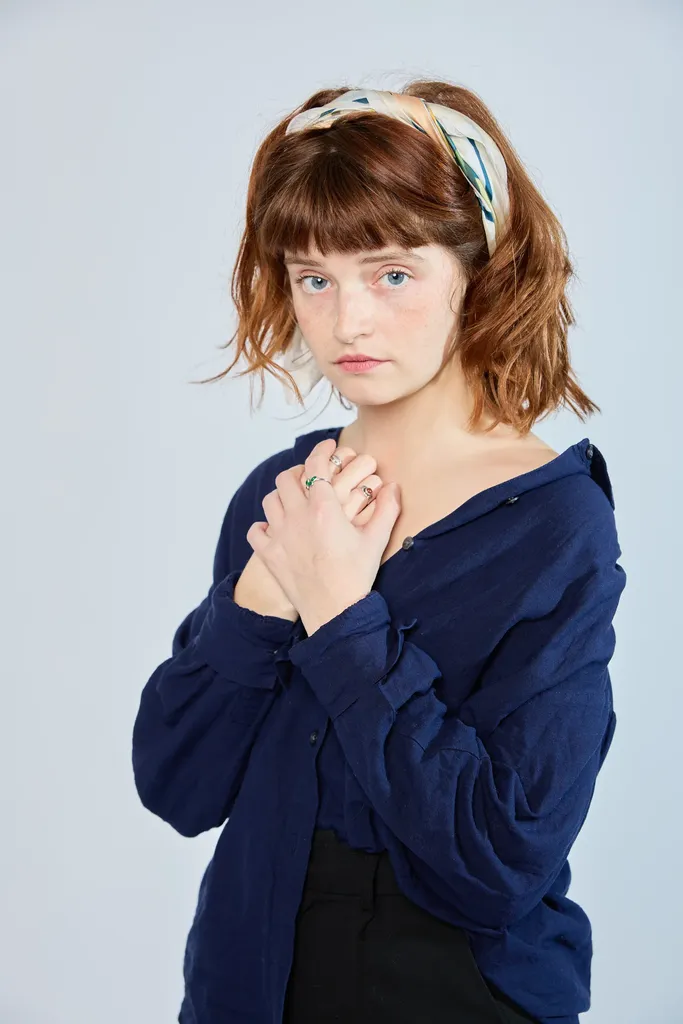 A red-haired woman with a scarf in her hair, hands crossed on her chest, grey-blue ground.
