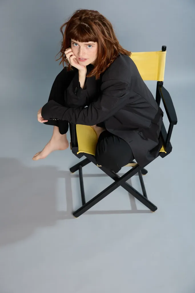 A red-haired woman sitting on a director’s chair with a yellow canvas, chin in hand, grey-blue ground.