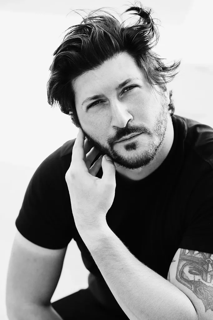 A bearded man in a black t-shirt, hand near his face, close portrait in black and white.