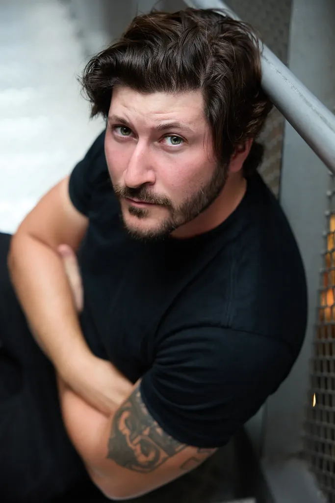 A bearded man in a black t-shirt, arms folded by a railing, seen from above, colour.