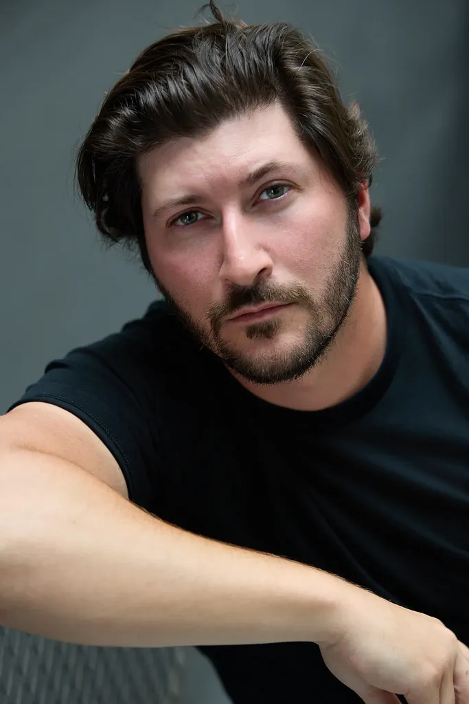 A bearded man in a black t-shirt leaning on his arm, looking straight at the camera, dark grey ground, colour.