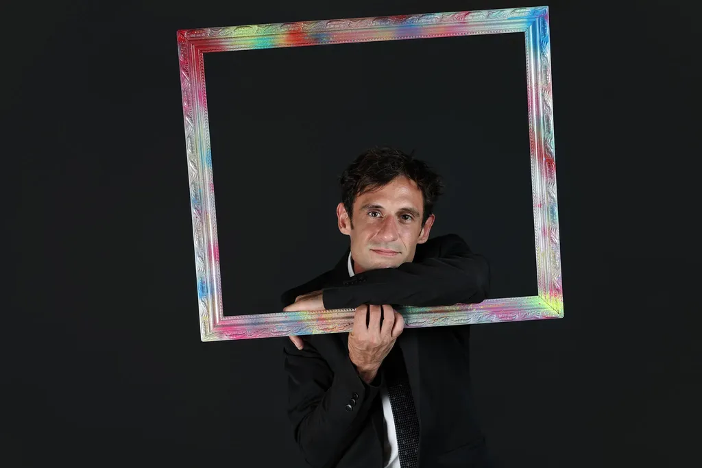 A man in a dark suit holding a multicoloured frame in front of him on a black ground, horizontal frame.
