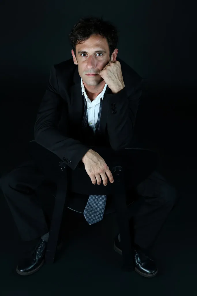 A man in a dark suit sitting with his chin in his hand on a black ground.
