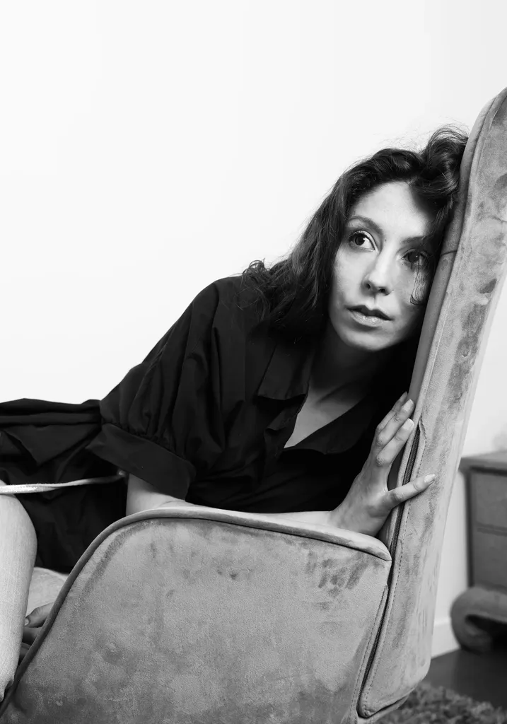A dark-haired woman dressed in black leaning against the back of a velvet armchair, in black and white.