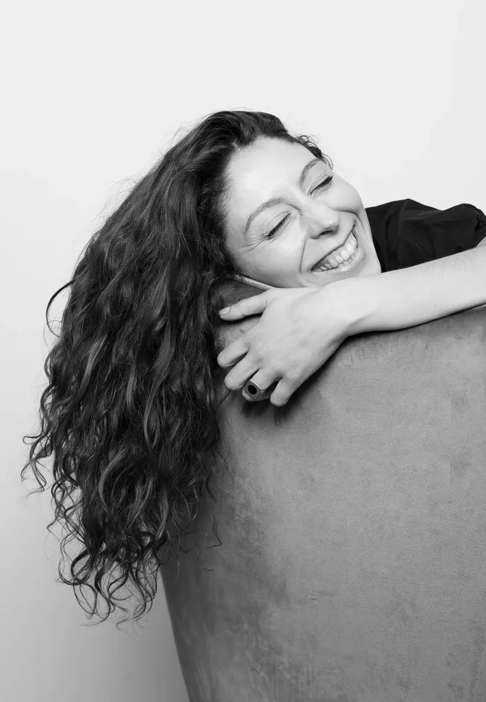 A woman with curly hair laughing, arms resting on the back of an armchair, in black and white.