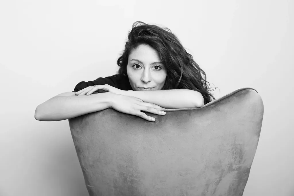 A woman with curly hair resting her chin on her folded arms over an armchair, horizontal frame, in black and white.