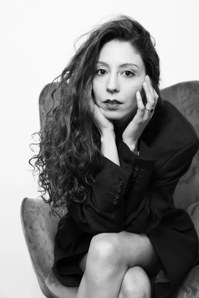 A woman with curly hair sitting in an armchair, hands near her face, in black and white.