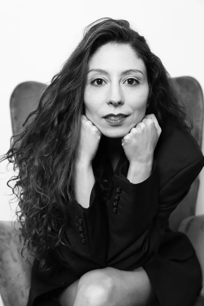 A woman with curly hair sitting in an armchair, direct gaze, fists under her chin, in black and white.