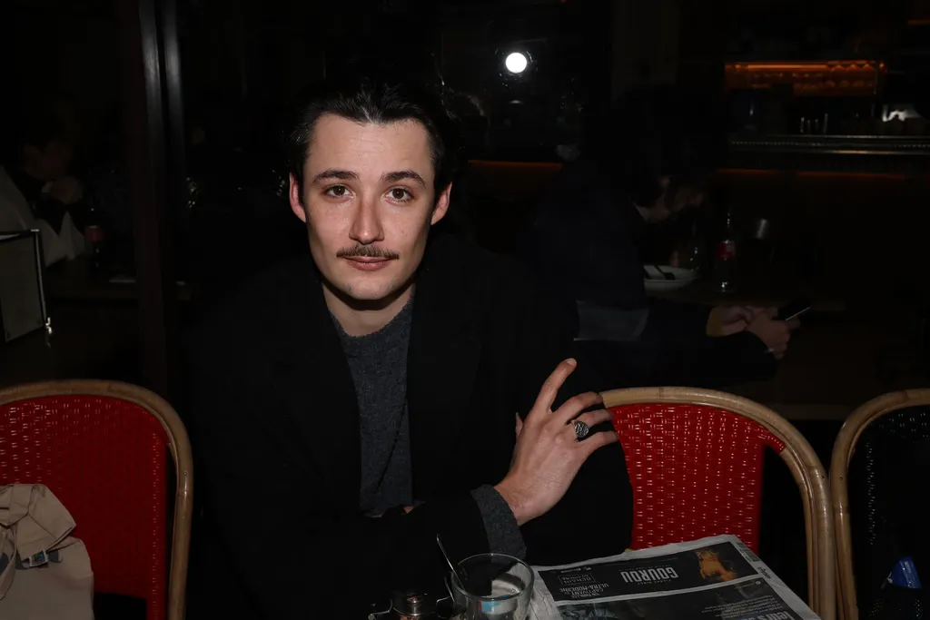 A young man with a moustache sitting at a café terrace in the evening between red chairs, horizontal frame.