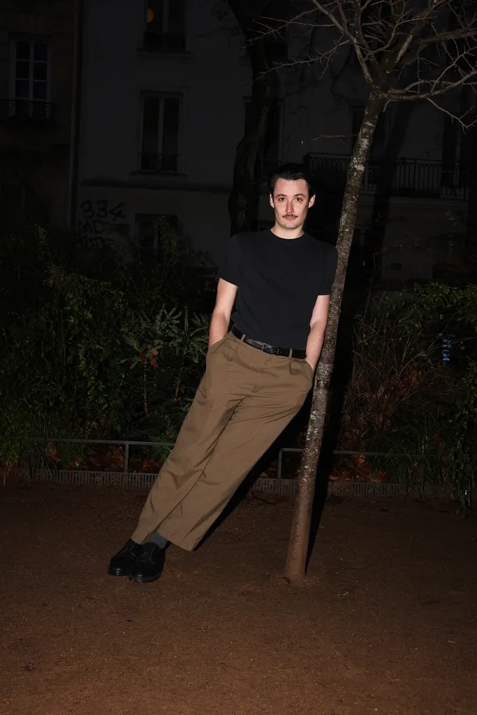 A young man in a black t-shirt leaning against a tree at night, buildings behind him.