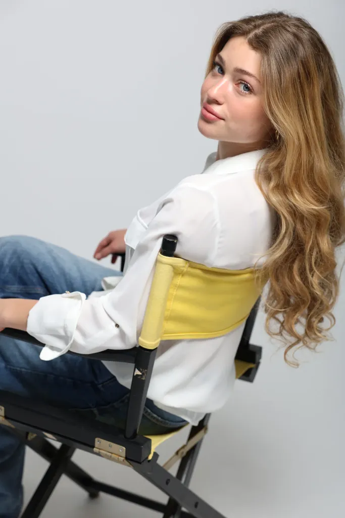 A young woman in a white shirt sitting sideways on a director’s chair with a yellow canvas, looking at the camera.