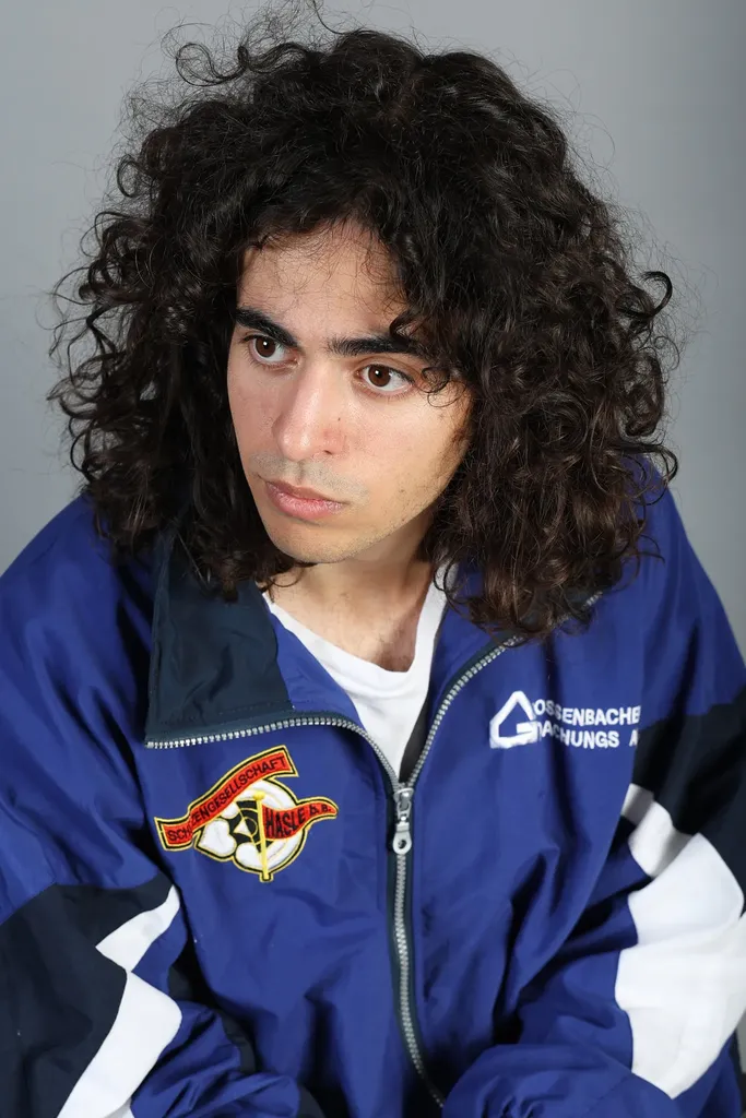 Portrait of a young man with curly hair in a blue track jacket, grey ground.