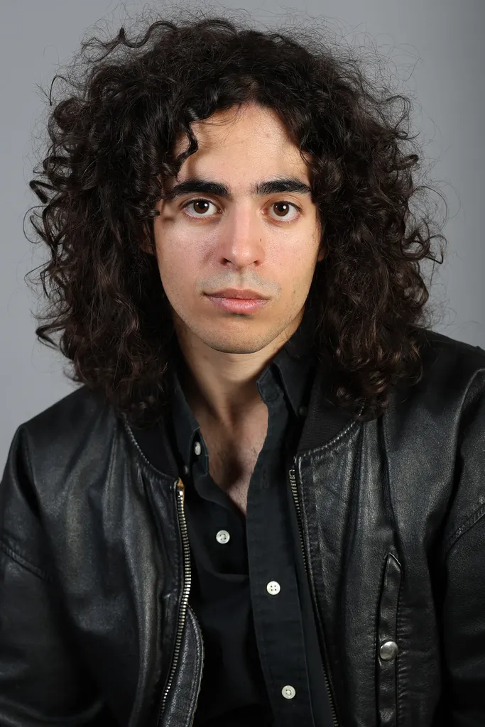 Front portrait of a young man with curly hair in a black leather jacket, grey ground.