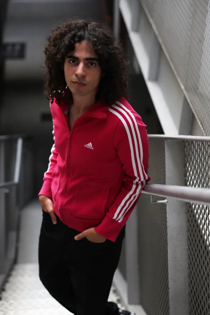 A young man with curly hair in a pink jacket, hands in his pockets, on a metal staircase.