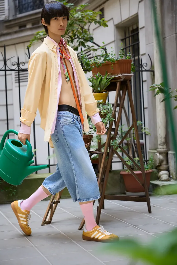 Young man with black hair in an open yellow shirt and cropped jeans, holding a green watering can, walking past potted plants in a courtyard, in colour.