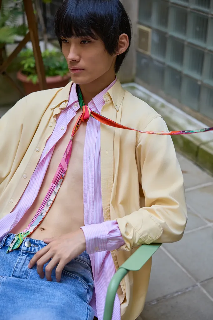 The same young man seated on a green chair, yellow shirt over a pink striped shirt and a colourful scarf, framed close in front of glass bricks, in colour.