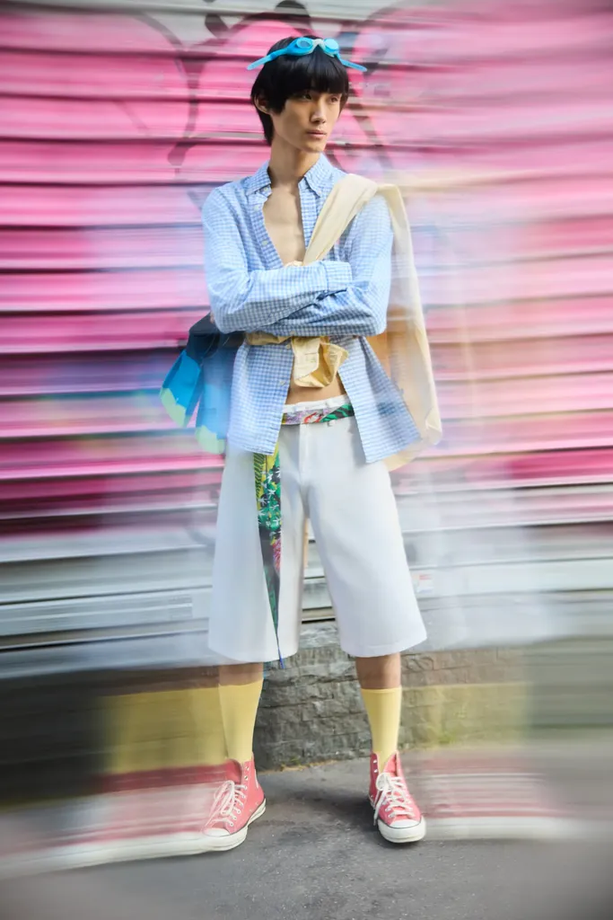 The same young man standing before a pink tagged metal shutter, blue shirt, yellow sweater over his shoulders and pink sneakers, the background blurred by motion, in colour.