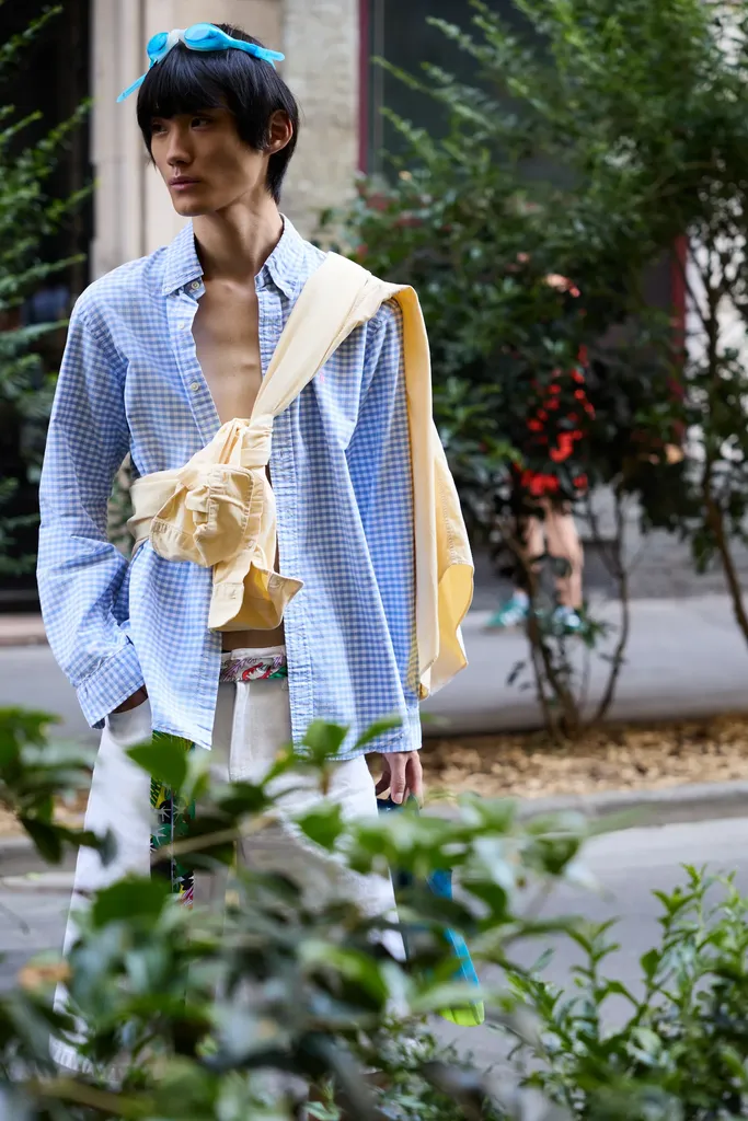 The same young man in a blue gingham shirt with a yellow sweater tied at the waist, surrounded by foliage in a street, in colour.