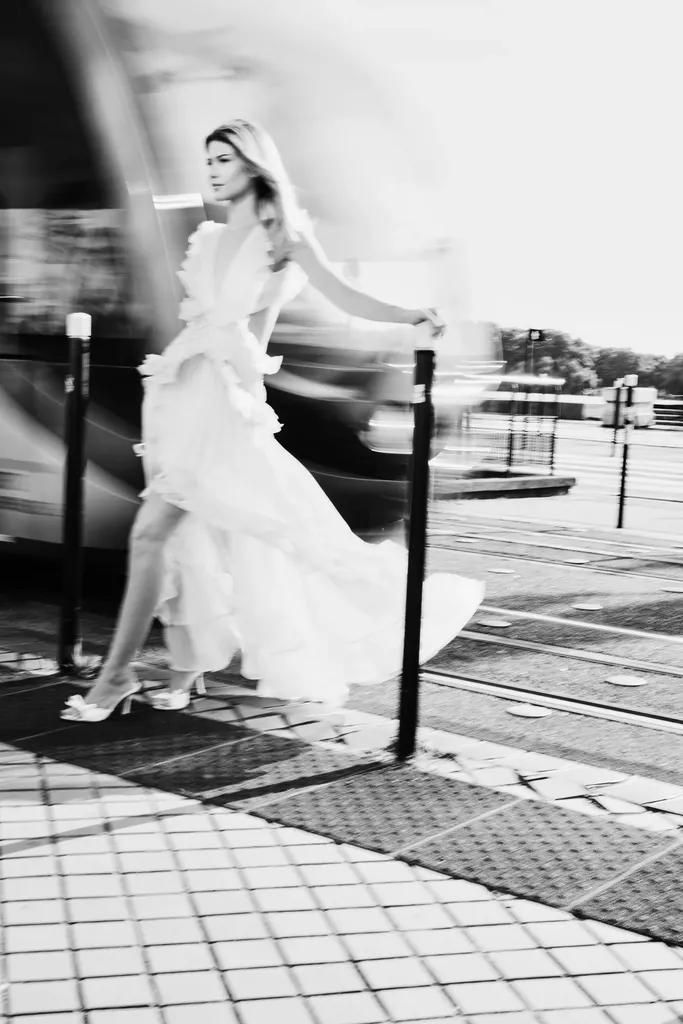 Woman in a long white ruffled dress walking on paving stones, the background blurred by motion, in black and white.