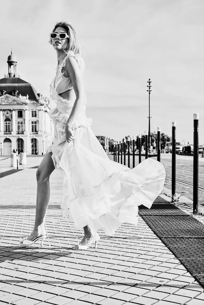 The same woman in a white ruffled dress and white sunglasses, the dress lifted by her stride, on a paved quay lined with bollards, in black and white.