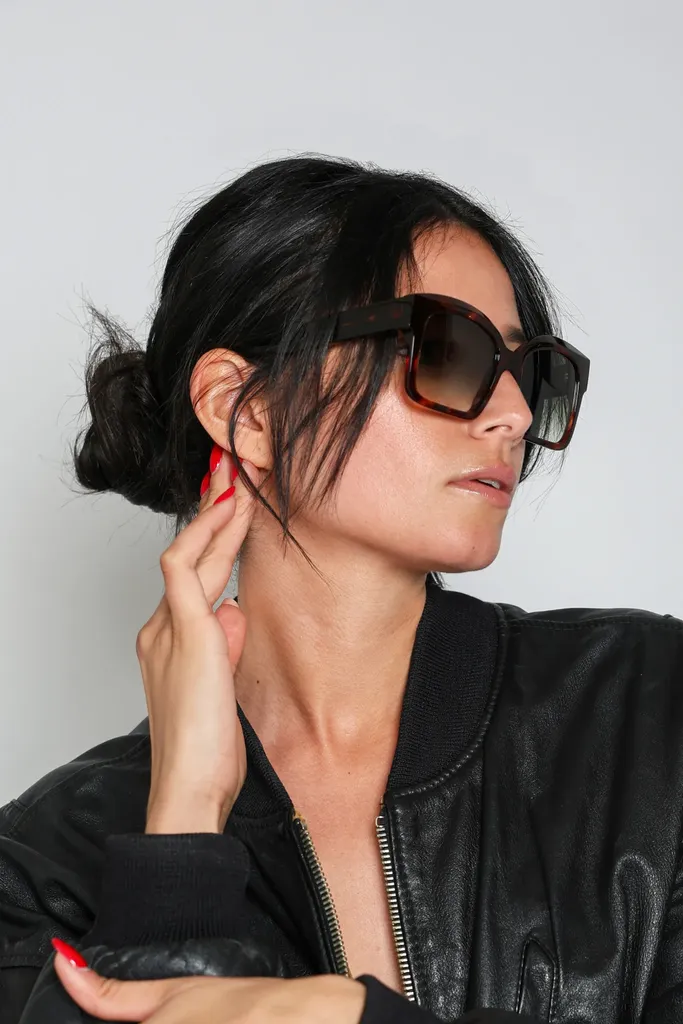 Close portrait of a dark-haired woman in large sunglasses and a black leather jacket, one hand at her ear, on a grey studio background, in colour.