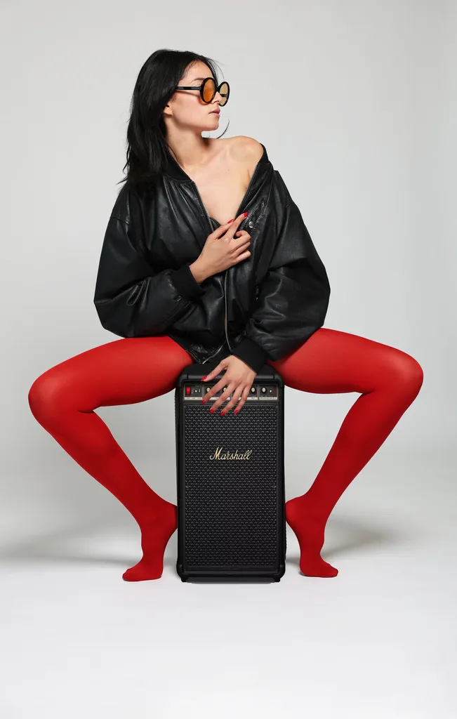 The same woman in a black leather jacket and red tights, seated on a black amplifier, round orange sunglasses, on a light studio background, in colour.