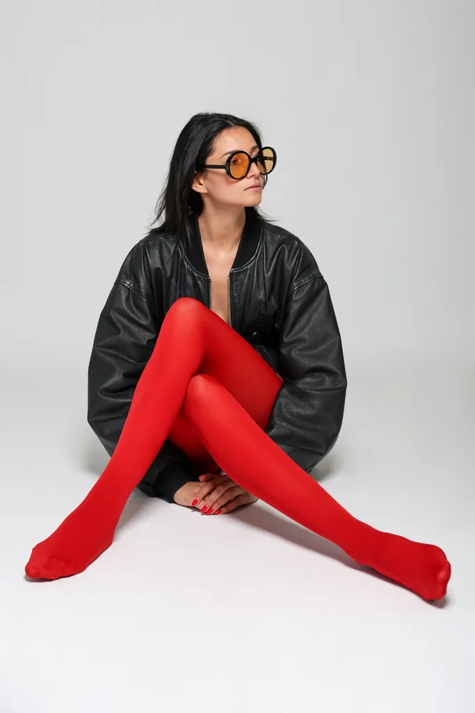 The same woman sitting on the floor, legs in red tights stretched out, black leather jacket, on a light studio background, in colour.