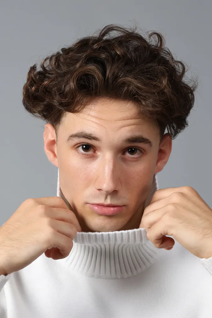 Close-up of a young man with curly hair pulling up the collar of a white turtleneck, on a grey background, in colour.