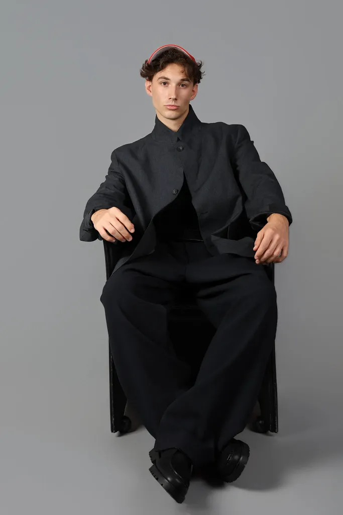 The same young man seated in a black suit and a red cap, full length on a grey studio background, in colour.