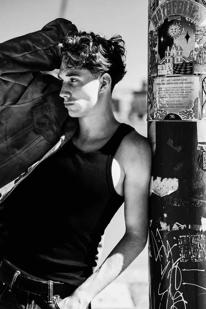 Young man in a black tank top and leather jacket leaning against a pole covered in posters, in full sun, in high-contrast black and white.