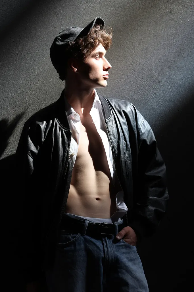 Young man in a cap and an open leather jacket, bare-chested, in hard side light against a dark wall, in colour.