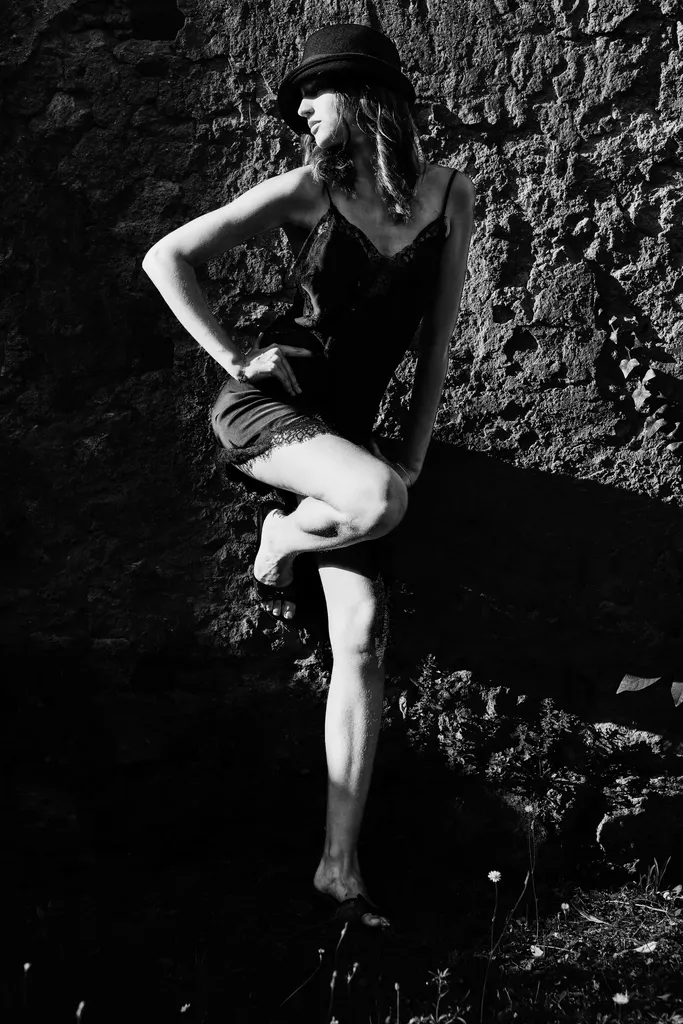 Woman in a black slip dress and hat, one knee raised against a rough stone wall, in full sun, in high-contrast black and white.