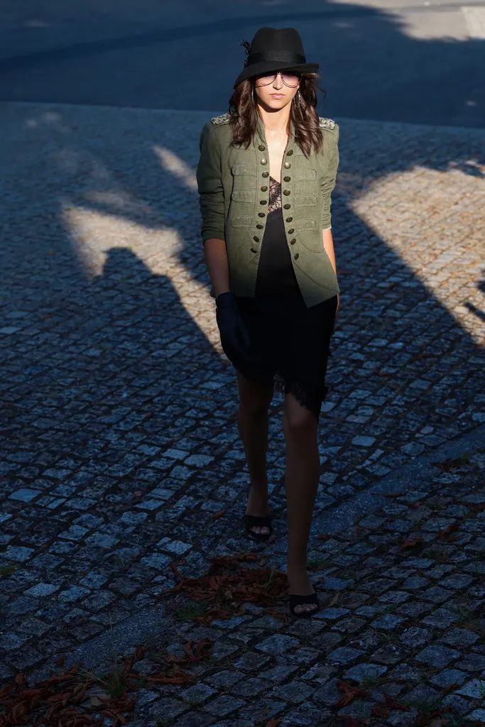 Woman in a black hat, khaki jacket with gold buttons and black dress walking on cobblestones in low light with long shadows, in colour.