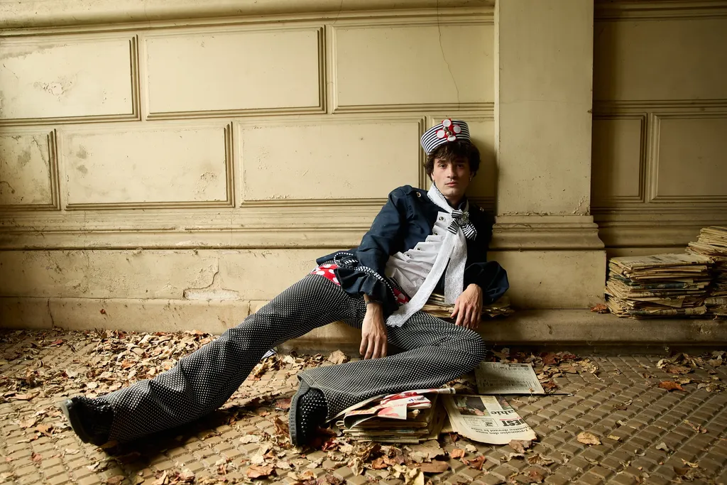Young man in a navy jacket and striped trousers sitting on the ground against a pale stone wall, among dead leaves and newspapers, in colour.
