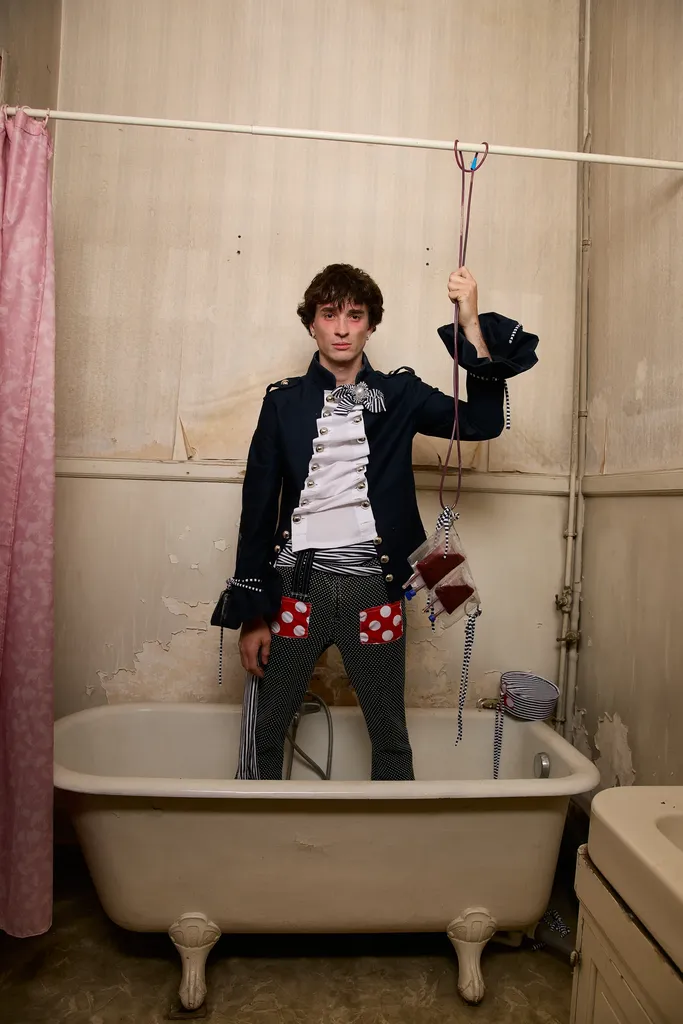 The same young man in a navy jacket with a white front, standing in an old bathtub in a worn bathroom, in colour.