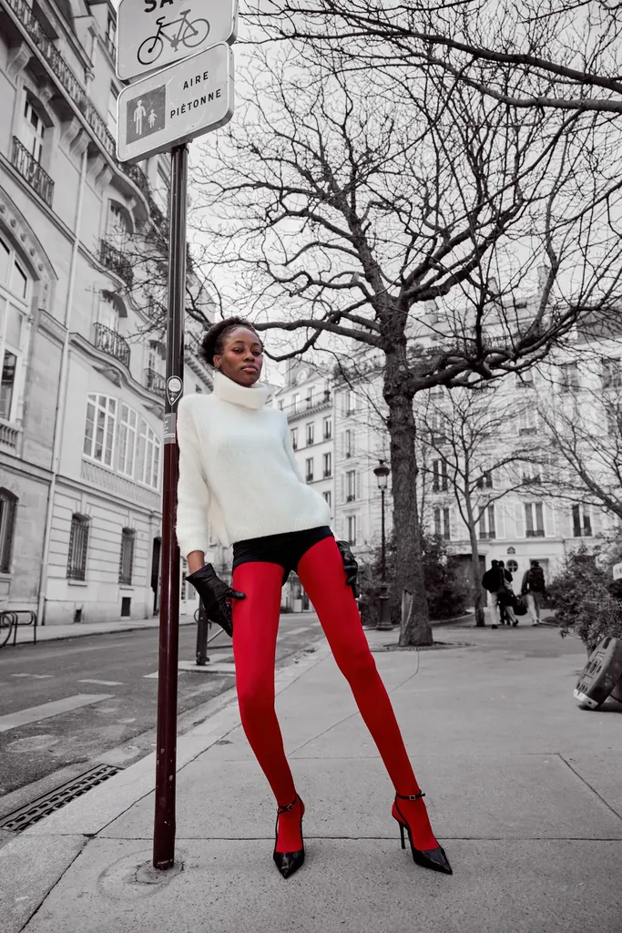 Woman in a white turtleneck and red tights leaning on a pole on a pavement, the street in black and white and the tights in colour.