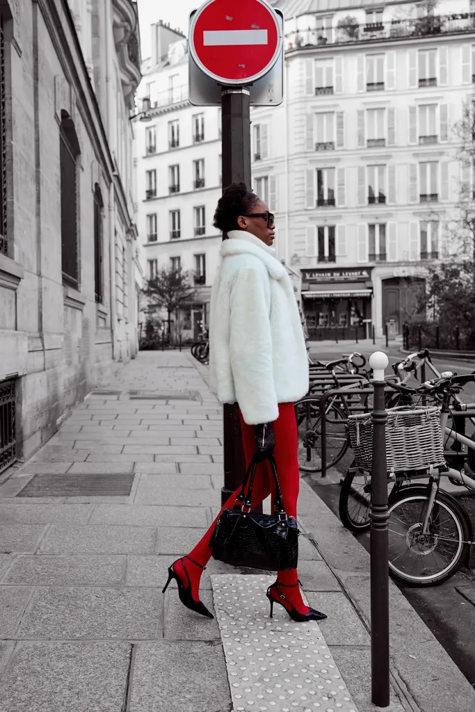 Woman in a white fur coat, dark sunglasses and red tights on a pavement near bicycles, the street in black and white and the tights in colour.
