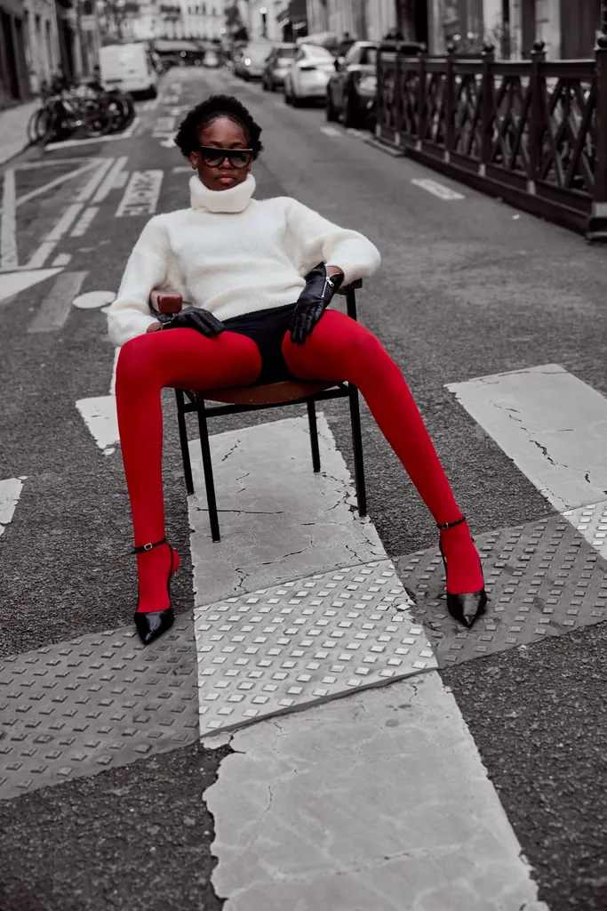 Woman seated on a chair in the middle of a pedestrian crossing, white sweater, black gloves and red tights, the street in black and white and the tights in colour.