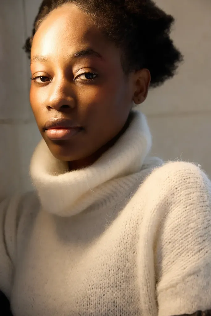 Close portrait of a young woman in a cream turtleneck, lit by a warm side light, in colour.