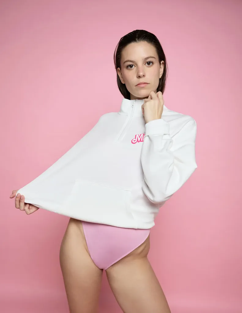 Woman with short hair in a white sweatshirt and pink bodysuit, holding the hem of her sweatshirt, on a pink paper backdrop, in colour.