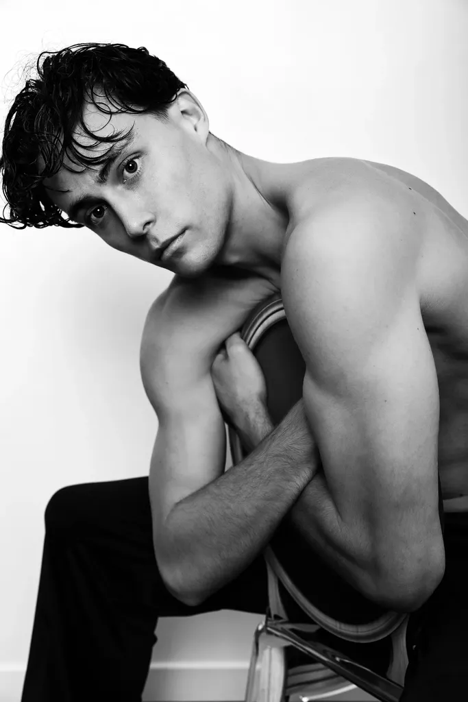 Bare-chested young man with curly brown hair, arms folded over the back of a chair, looking into the lens, in black and white.