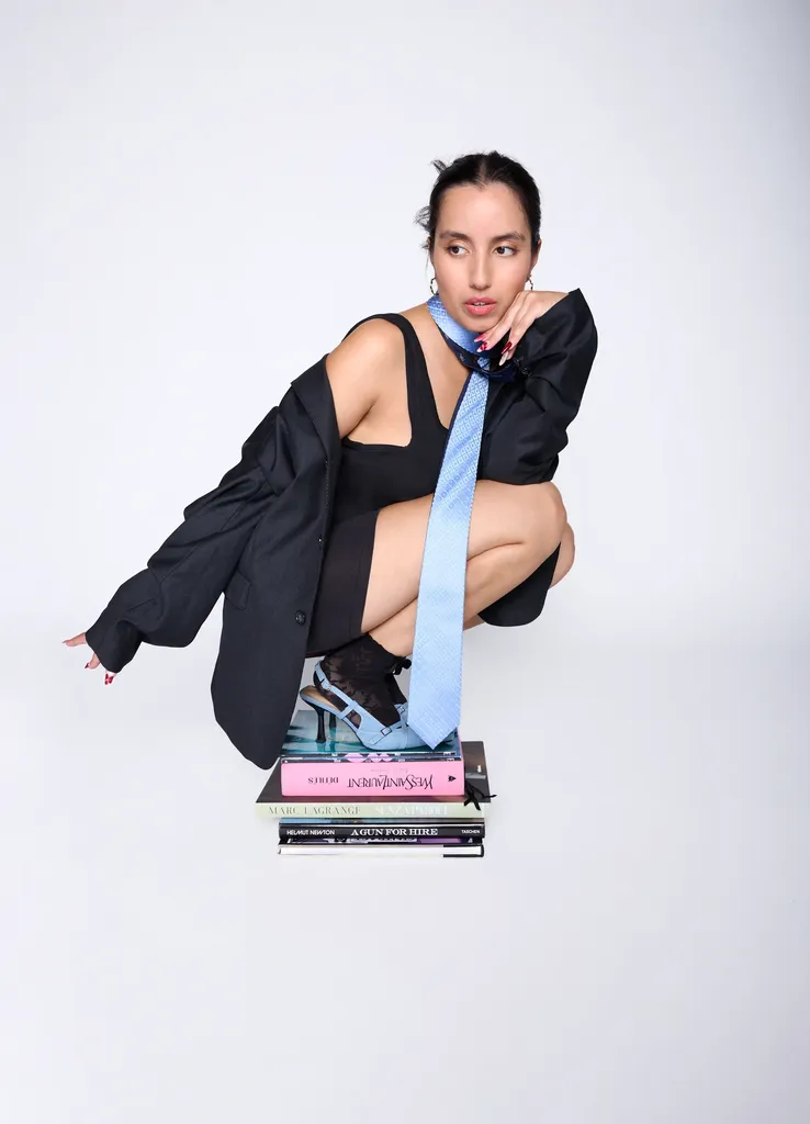 Woman crouching on a stack of books, oversized black blazer and pale blue tie, on a light grey background, in colour.
