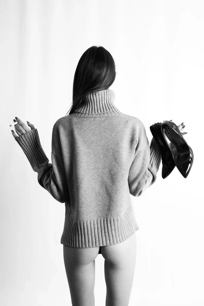 Woman seen from behind in a turtleneck sweater, holding a pair of black heels, on a white background, in black and white.