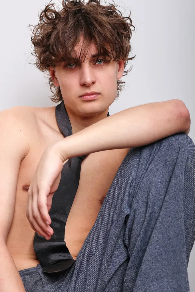 Young man with tousled brown hair, bare-chested with a dark tie, his arm resting on a raised knee, on a light background, in colour.